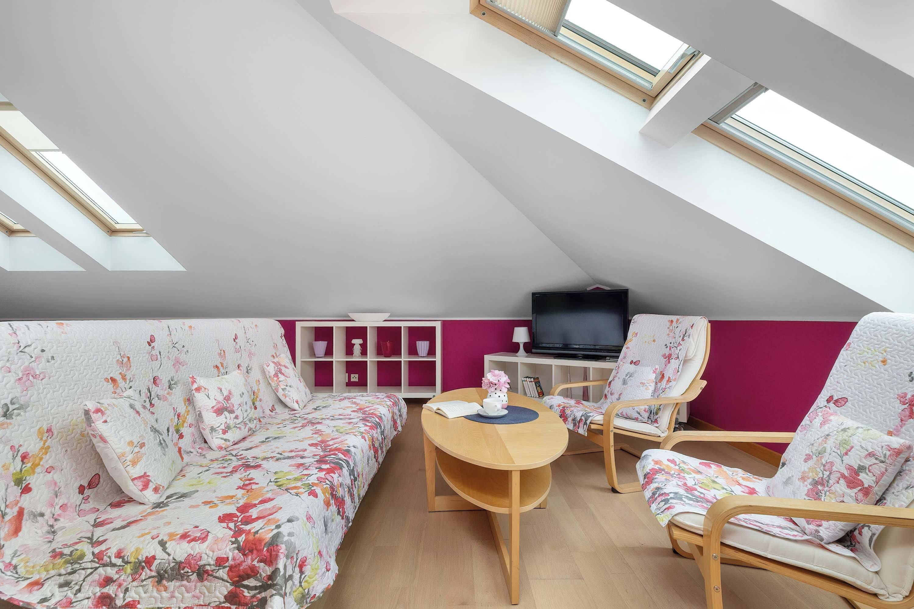 Ein modernes Schlafzimmer im Loft-Stil mit schrägen Wänden, Dachfenstern, einem Bett mit floralem Bettzeug, zwei Stühlen, einem kleinen Tisch und einem Fernseher.