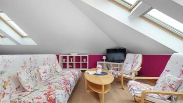 A modern loft bedroom with a sloped ceiling, skylights, a bed with floral bedding, two chairs, a small table, and a TV.