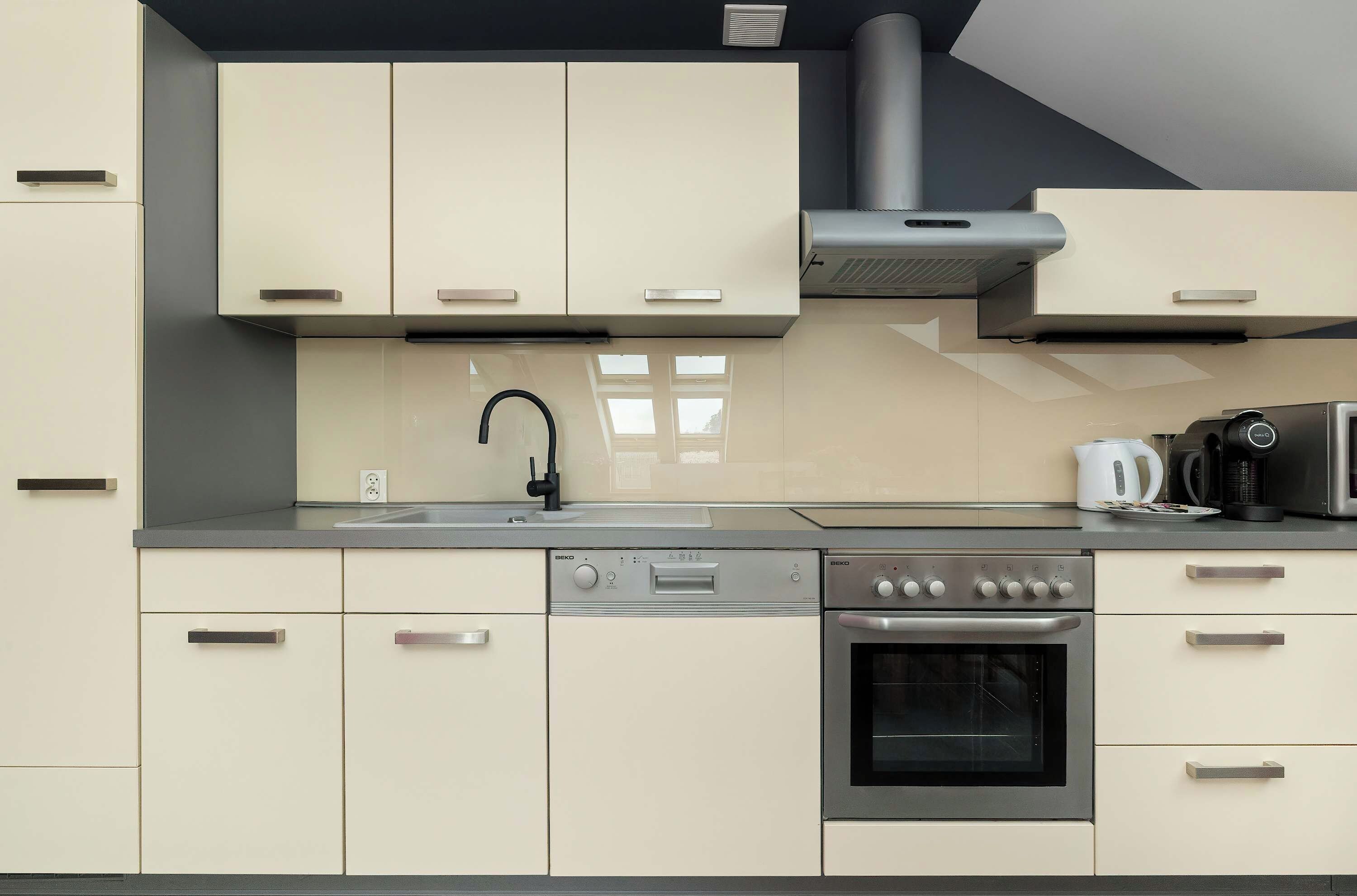 A modern kitchenette with white cabinets, a sink, an induction cooktop, and a built-in oven. The kitchen has a light countertop and a minimalist design.