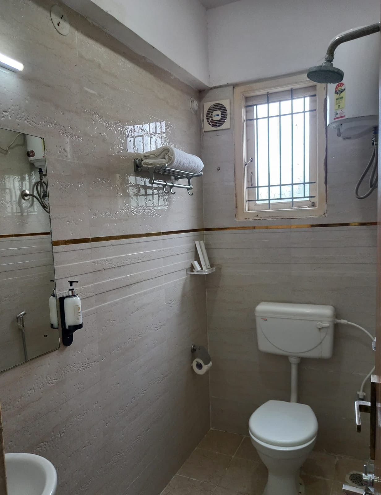 A small bathroom with a toilet, a shower, a window with bars, a towel rack, and a mirror.