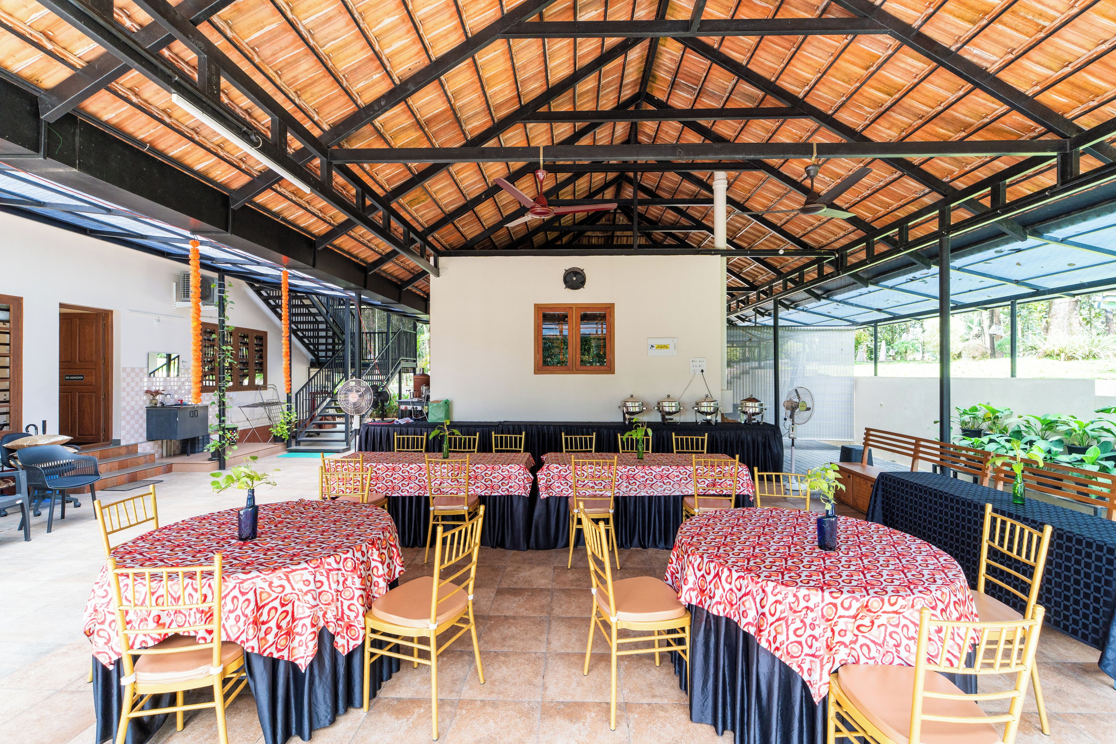Un espacio de comedor al aire libre con techo, varias mesas y sillas, una escalera y vistas del entorno verde.