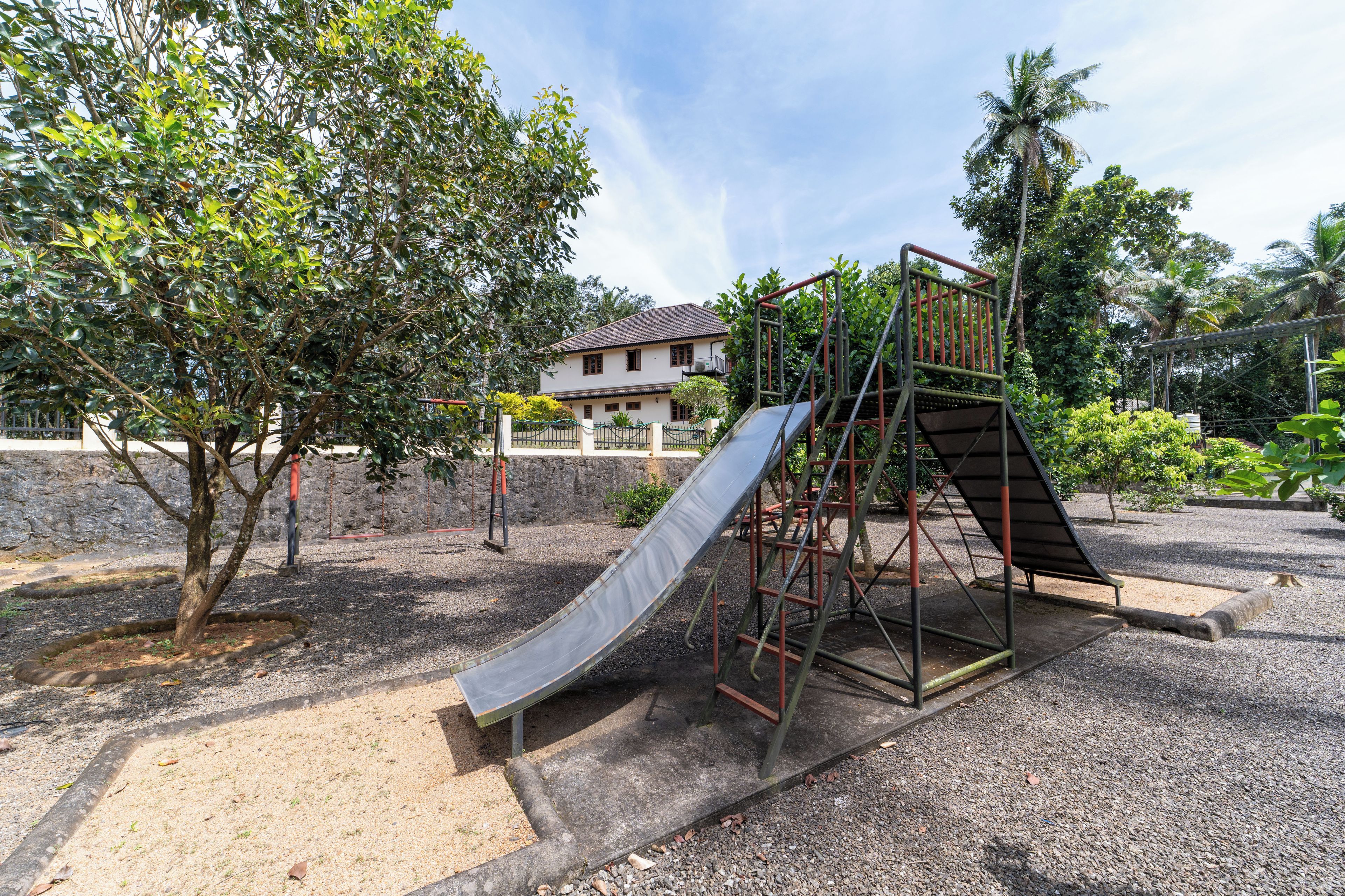 Un parque infantil con tobogán y estructura para escalar, rodeado de árboles y una casa al fondo.