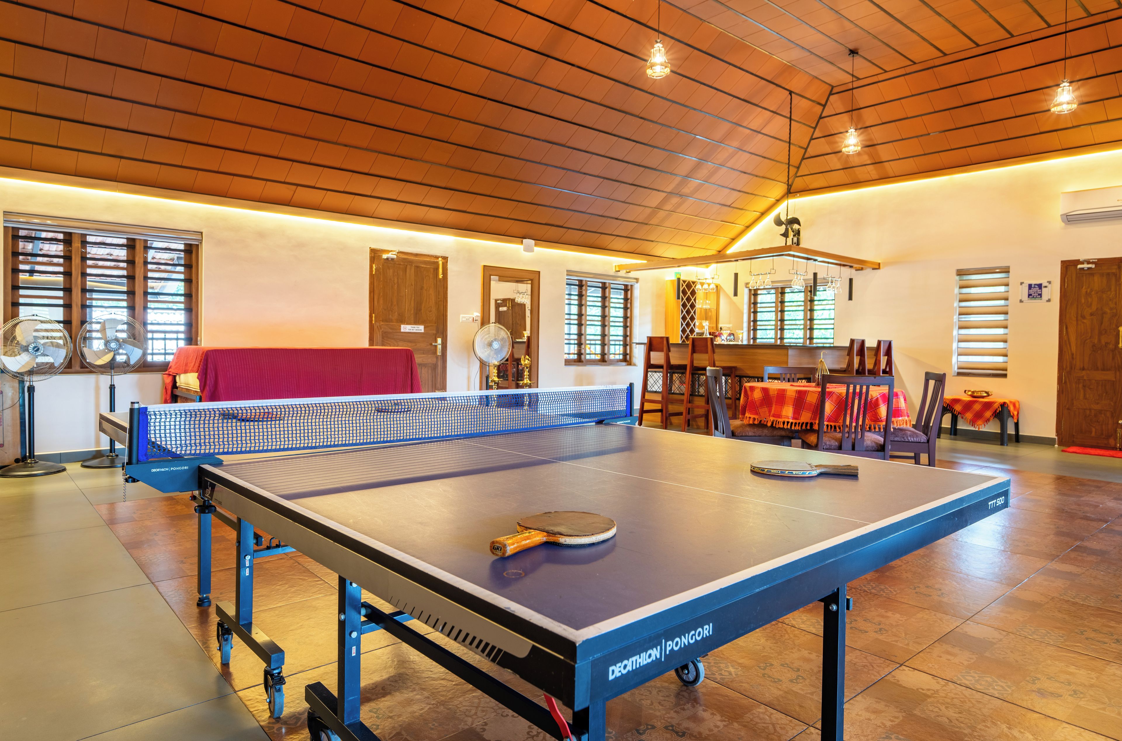 A ping pong table with a net, a table tennis paddle, and a table tennis ball in a room with wooden flooring, a high ceiling, and a dining area in the background.