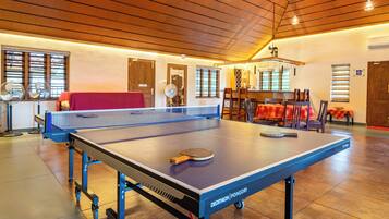 A ping pong table with a net, a table tennis paddle, and a table tennis ball in a room with wooden flooring, a high ceiling, and a dining area in the background.