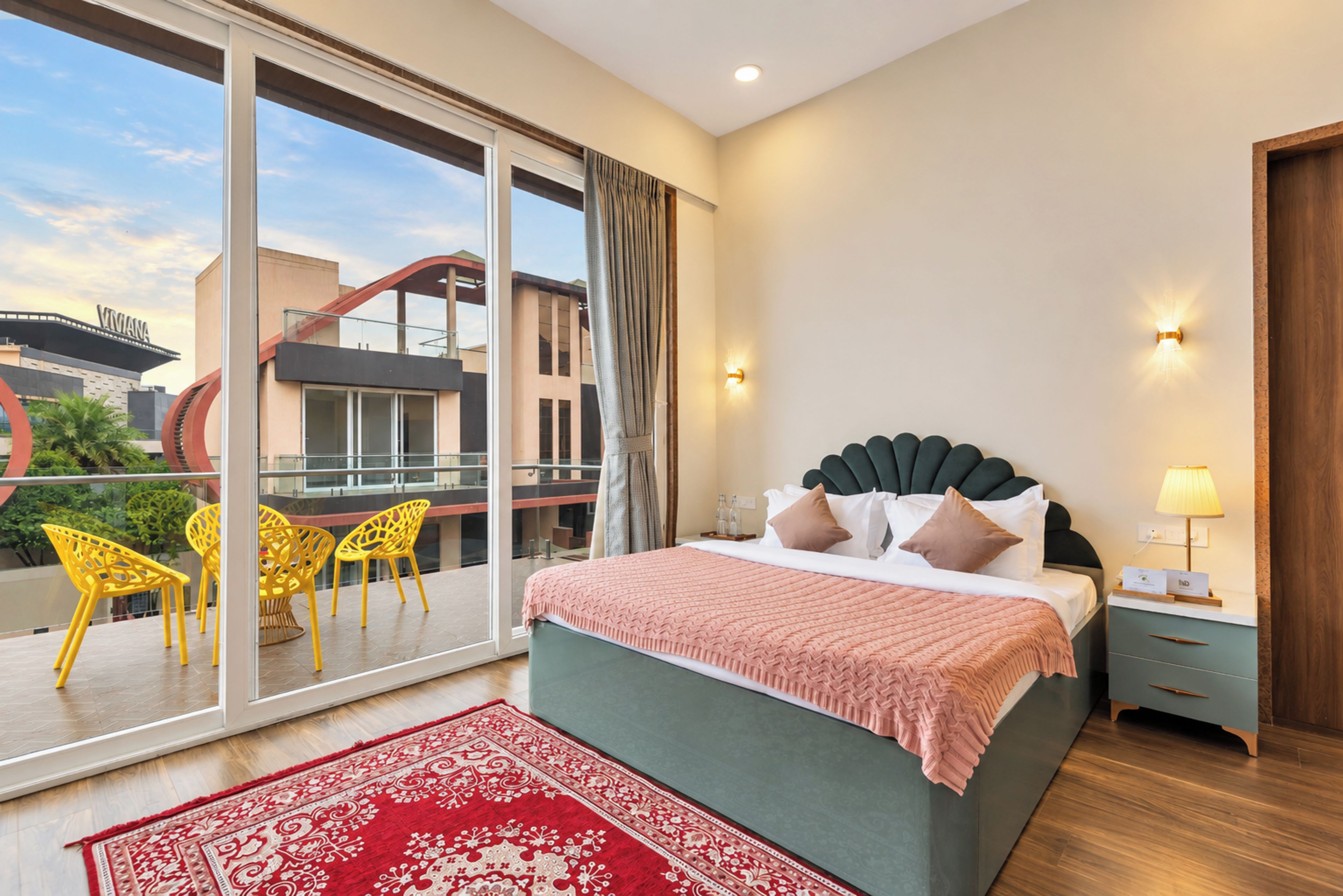 A hotel room with a large bed, a balcony with yellow chairs, and a view of a building with a red structure.