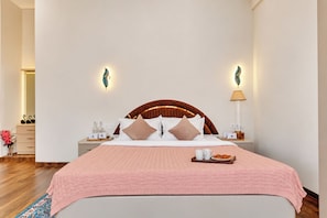 A hotel room with a large bed, a wooden headboard, two bedside tables with lamps, and a pink bedspread. - ELIVAAS Signature Begonia Paradise 3BHK (Varsoli)