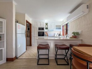 Eine moderne Küche mit weißen Schränken, einem Kühlschrank, einem Backofen, einer Mikrowelle und einem Spülbecken. Zwei Hocker und ein Holztisch sind ebenfalls vorhanden.