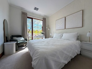 A bedroom with a large bed, a sitting area with a chair and a table, a window with curtains, and two wall-mounted artworks. - Dimora Court City Retreat (Adelaide)