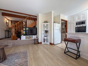 Ein modernes Wohnzimmer mit einer Holztreppe, einem Flachbildfernseher, einem weißen Schrank und einer Küchenzeile mit einem Doppelbackofen und einem Kühlschrank.