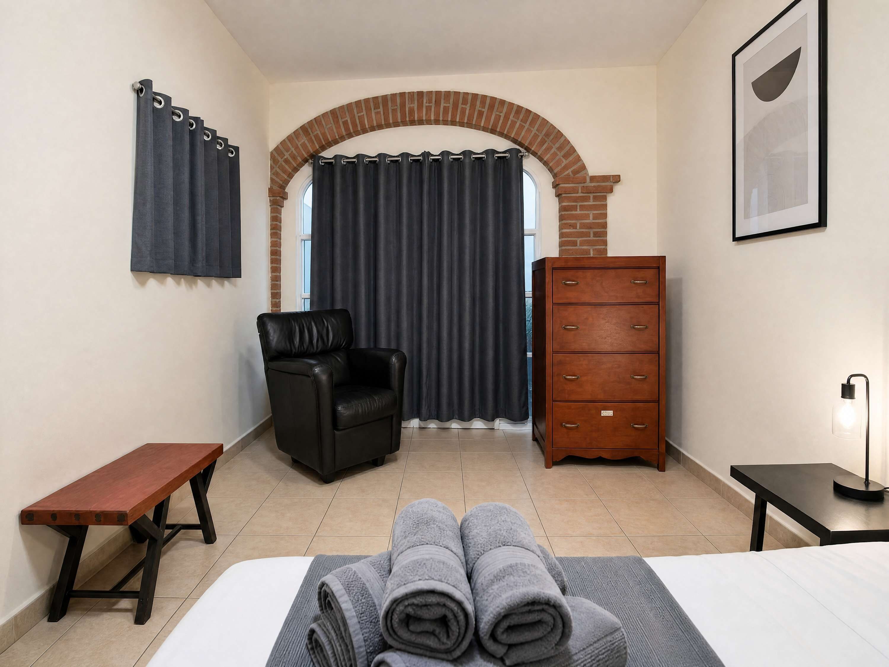 A hotel room with a bed, a bench, a chair, a dresser, and a desk.