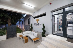 Entrada de hotel con mostrador de recepción, zona de estar con sofá y mesa de centro, y una escalera que lleva a la entrada.