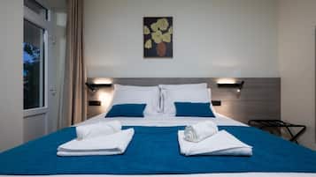 A hotel room with a bed, two pillows, a blue bedspread, two rolled towels, a painting on the wall, and a window with curtains.