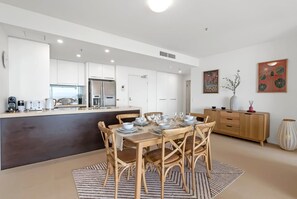 A modern kitchen with white cabinets, a central island with seating, and a dining area with a table set for four. - 79 Harbour Lights with Parkland & Marina Views (Cairns City)