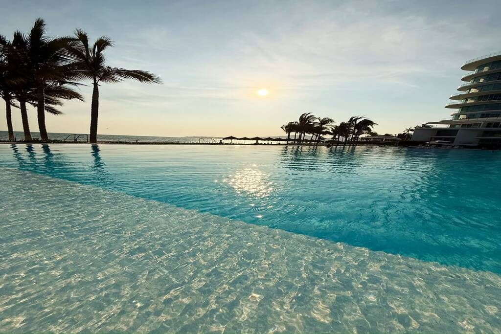 Une piscine aux eaux cristallines offrant une vue sur le coucher de soleil, les palmiers et le bâtiment de l’hôtel.