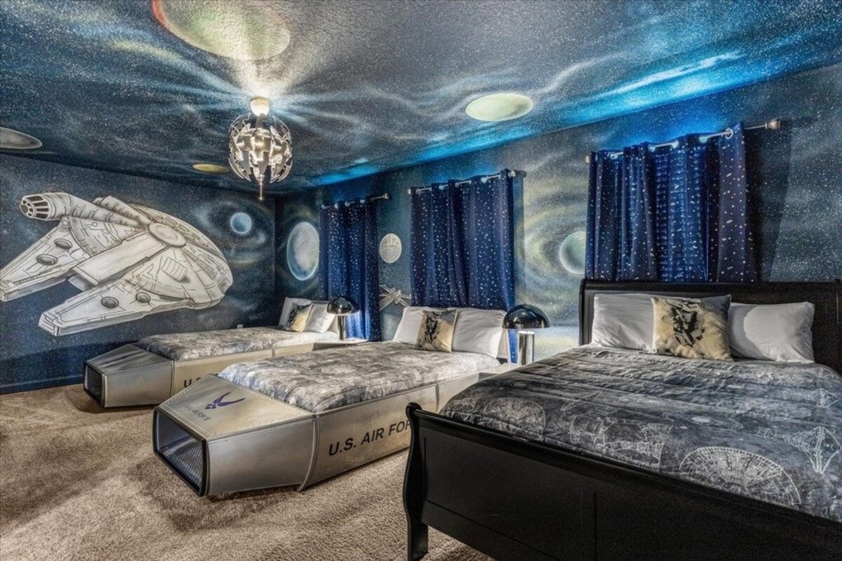 A bedroom with a space-themed mural, a large bed, two smaller beds, and a ceiling light fixture.