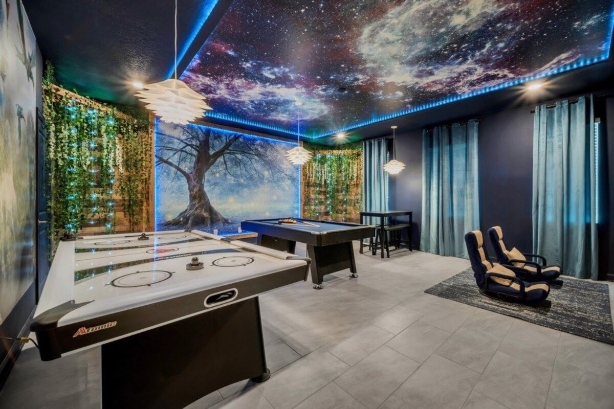 A room with a pool table, foosball table, and a pool table with a starry ceiling mural.