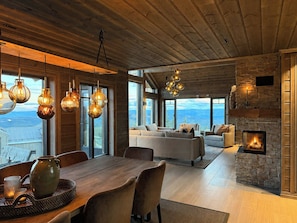 A spacious living area with a fireplace, large windows, and a dining table with chairs. - Family cabin by the ski trails on Sjusjøen (Sjusjøen)