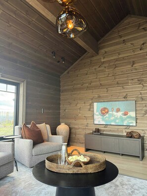 Ein modernes Wohnzimmer mit Holzbalkendecke, einem runden schwarzen Tisch mit einem gewebten Tablett, einem grauen Sofa und einem Gemälde an der Wand.