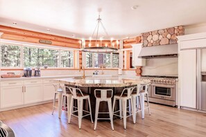 A spacious kitchen with a large island, white cabinetry, a stone accent wall, and a view of the forest through large windows. - New! Bearadise ~ Timberlodge Mountain Retreat (Big Bear Lake)