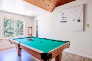 A pool table in a room with a large window, a framed drawing on the wall, and wooden flooring. - New! Bearadise ~ Timberlodge Mountain Retreat (Big Bear Lake)