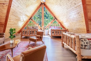 A cozy attic room with a high ceiling, wooden beams, a large window, and a view of greenery outside. - New! Bearadise ~ Timberlodge Mountain Retreat (Big Bear Lake)