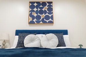 Une chambre à coucher avec une tête de lit bleue, des oreillers blancs à motifs, une couvre-lit bleue et une œuvre d’art abstraite accrochée au mur.