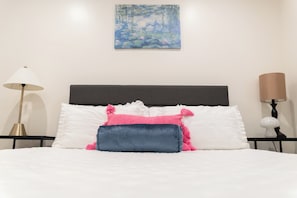 Een bed met witte beddengoed en decoratieve kussens, een schilderij erboven en nachtlampjes aan weerszijden.