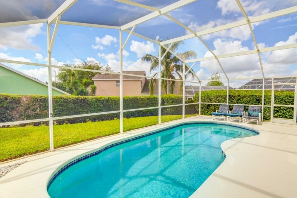 A swimming pool with a white deck, surrounded by a white fence, and a view of a residential area with houses and greenery.