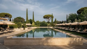 Una piscina rectangular amb gandules i ombrel·les a banda i banda, envoltada de vegetaci ó i arbres.