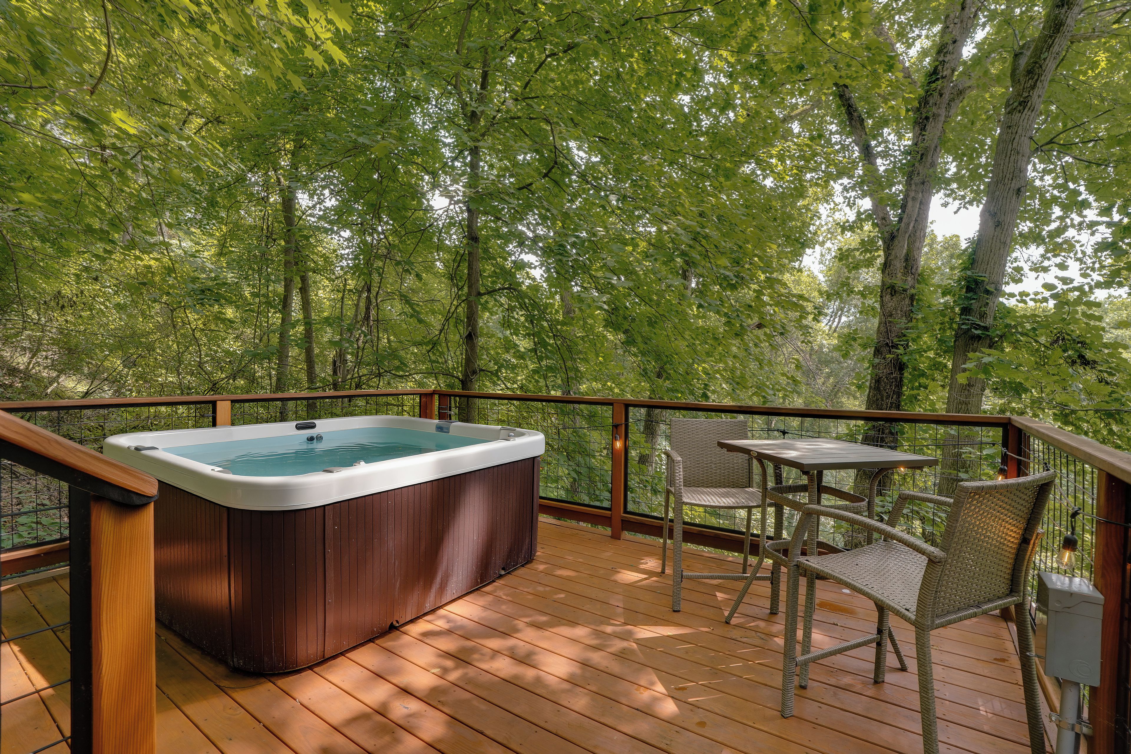 Een hottub op een houten terras met een tafel en stoelen, omgeven door een metalen reling en bomen.