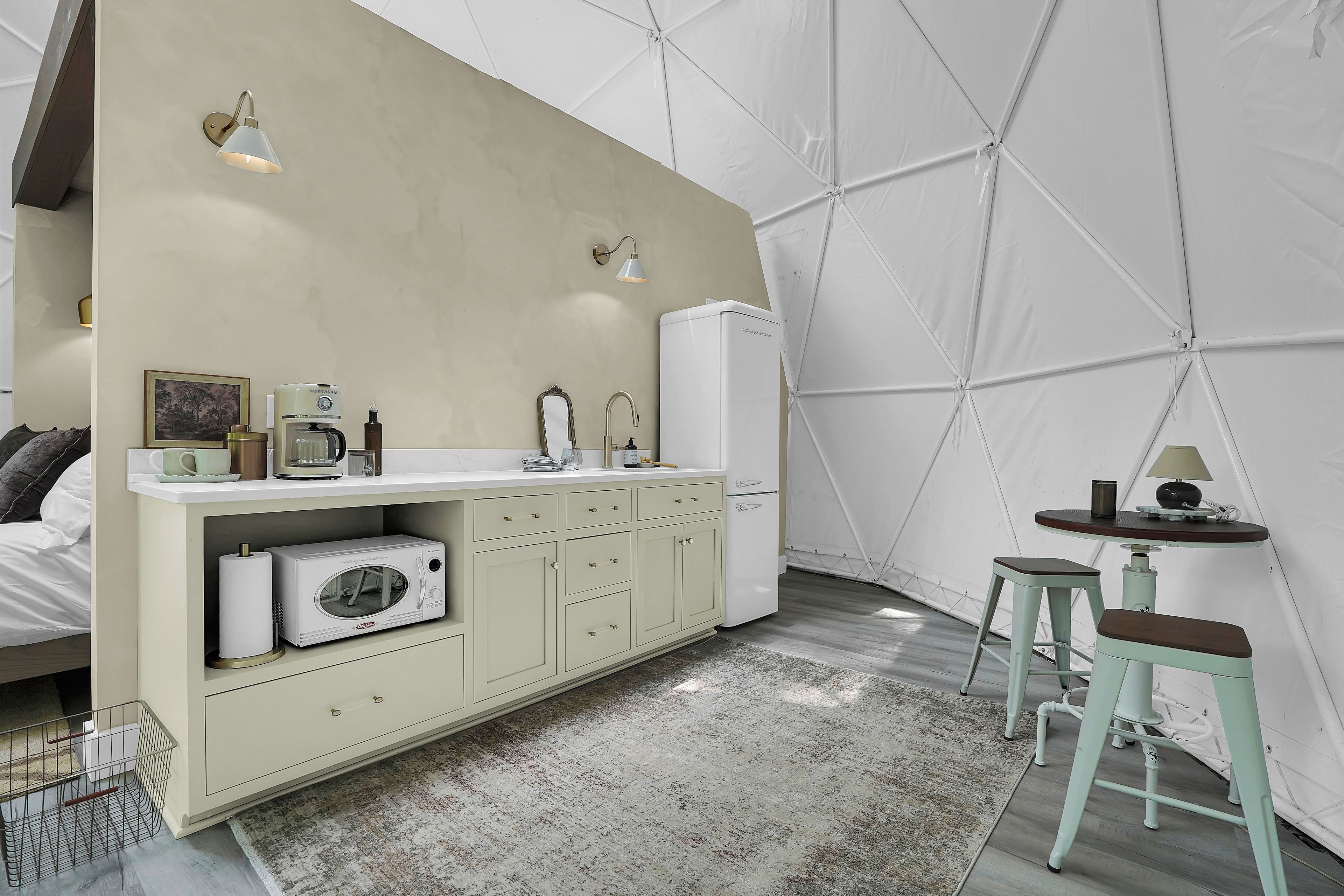 A modern bedroom with a bed, a built-in wardrobe, a small kitchenette, and a seating area with stools.