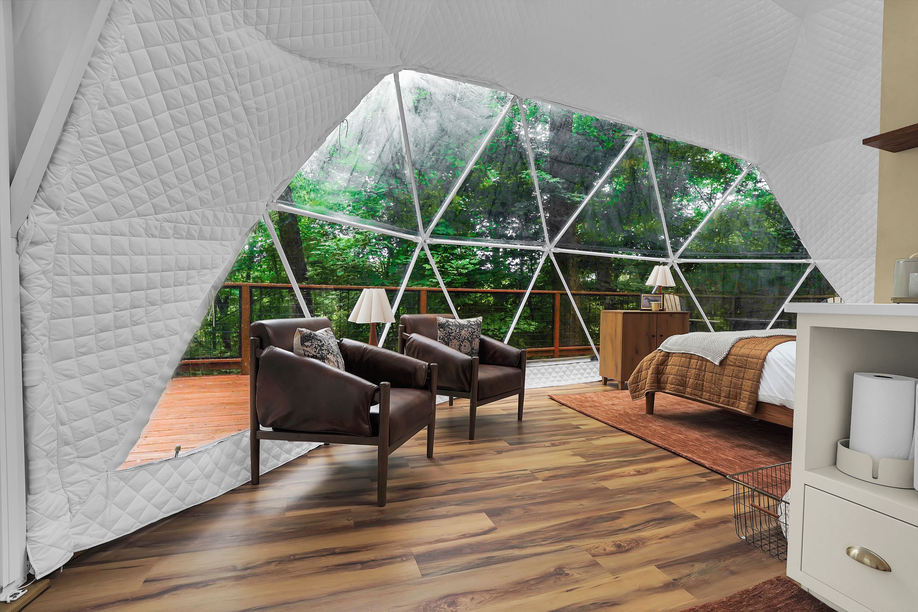 Una habitación con carpa, con cama, una silla, piso de madera y vista a un bosque a través de las paredes traslúcidas de la carpa.