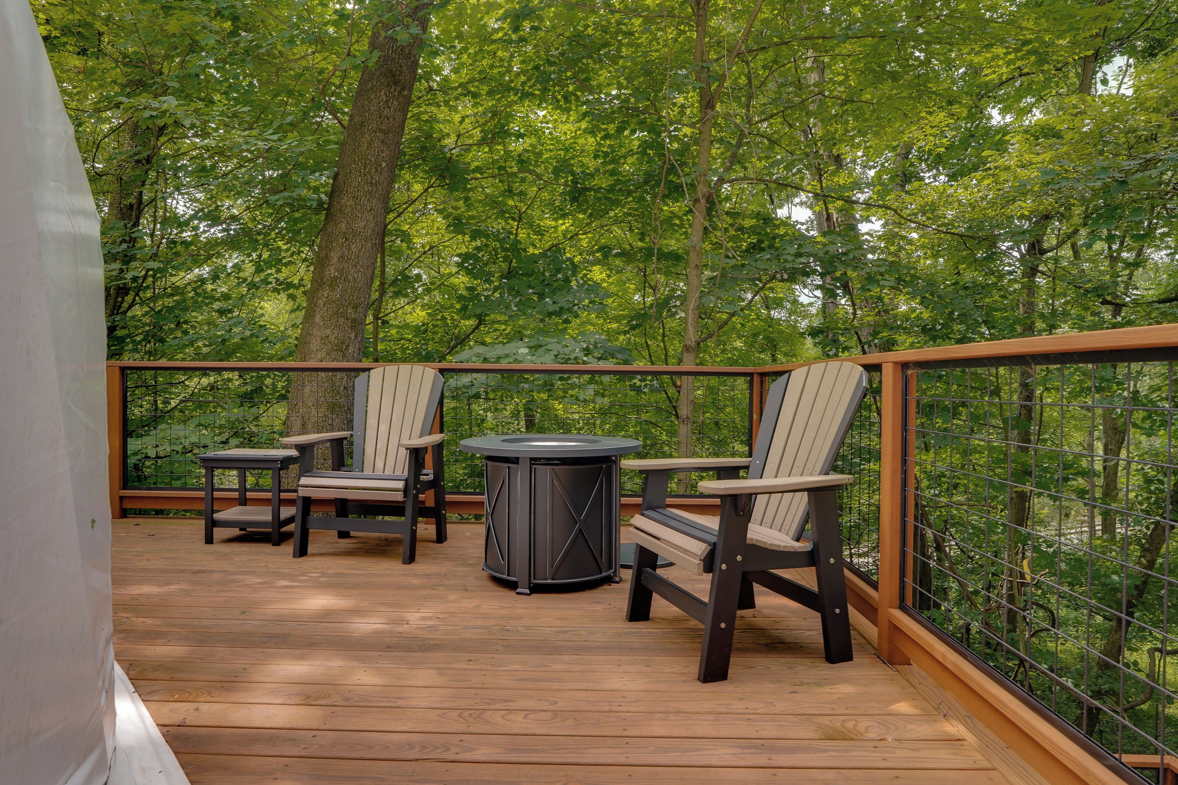 A wooden deck with outdoor furniture, including chairs and a table, surrounded by a metal railing and trees.