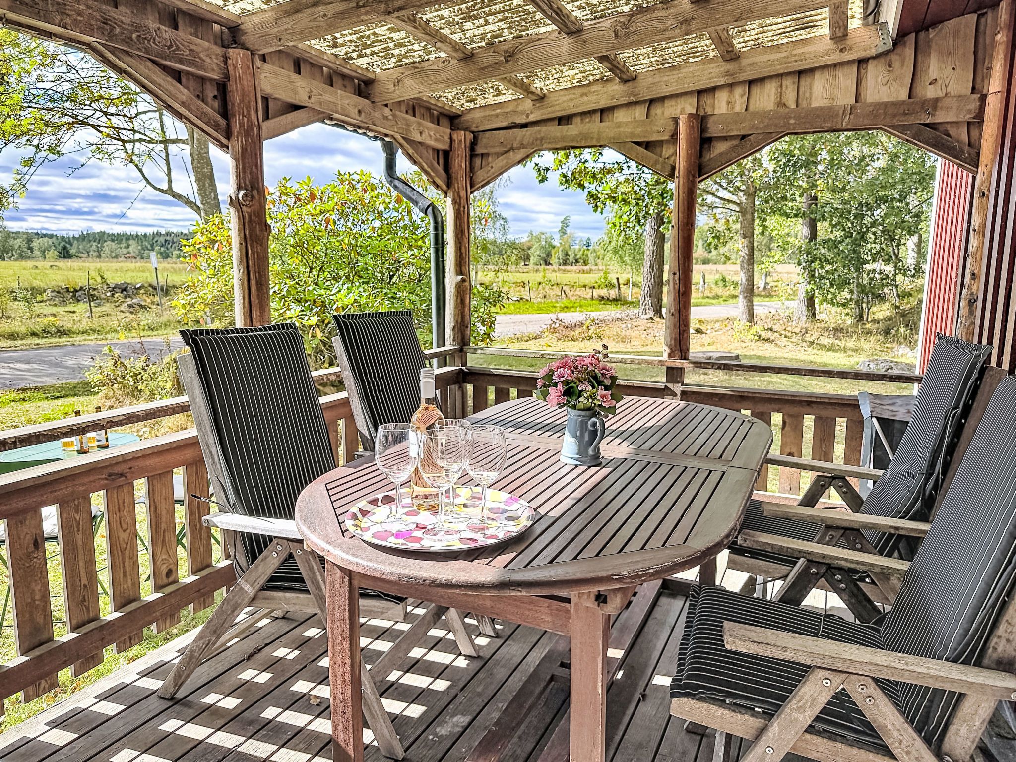 Um deck de madeira com uma mesa redonda e cadeiras, uma pérgula acima e vista para uma paisagem rural.