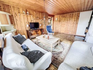 A cozy living room with wooden walls, a flat-screen TV, a blue armchair, and a white sofa with patterned cushions. - Tallstugan by Interhome (Kisa)