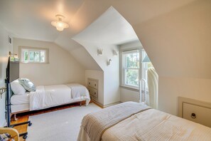 Ein Schlafzimmer mit schrägem Dach, einem Bett, einer Kommode und einem Fenster mit Blick auf Bäume.
