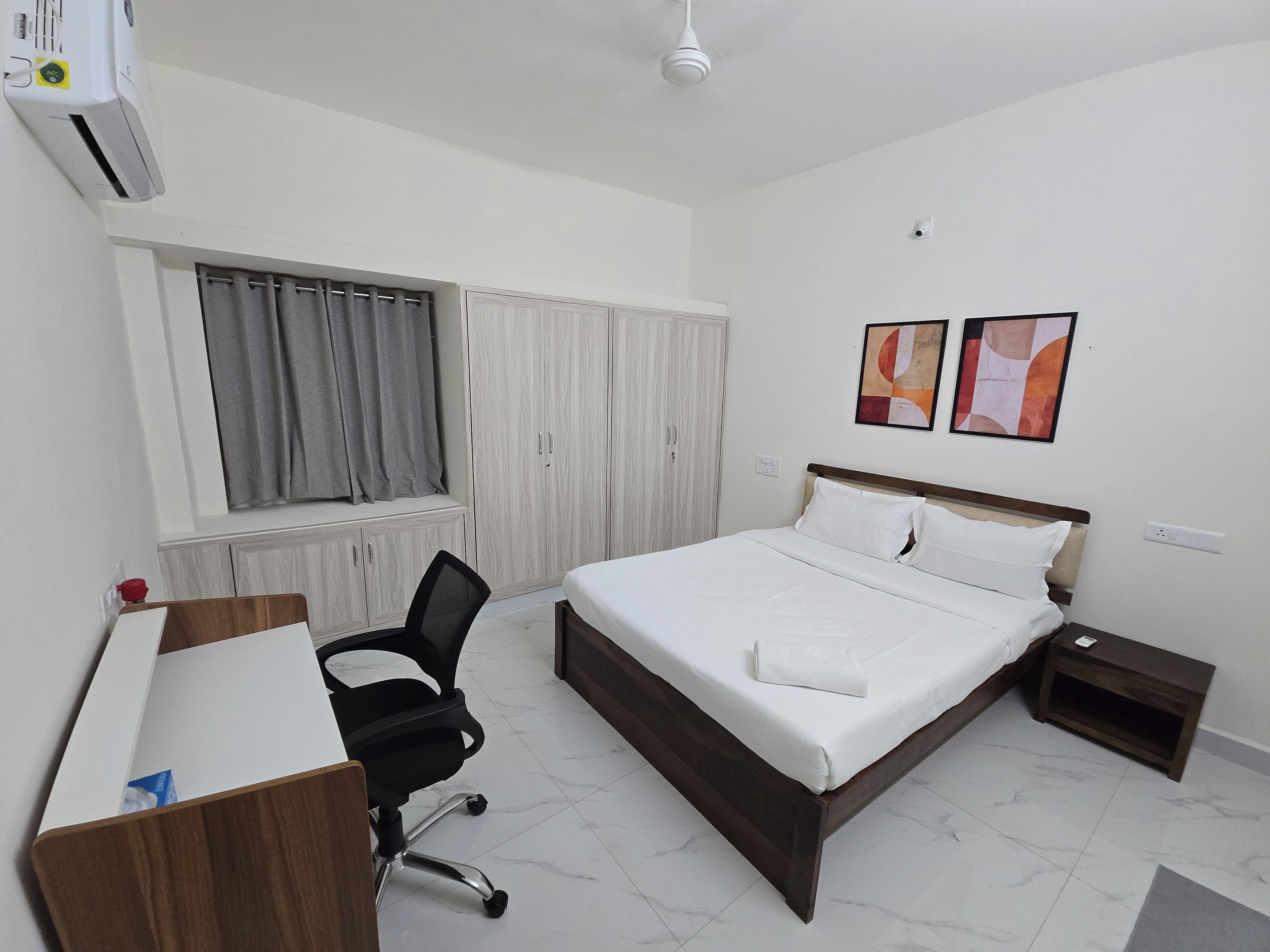Une chambre d’hôtel comprenant un lit, un bureau, une chaise, une fenêtre avec des rideaux et deux œuvres d’art accrochées au mur.
