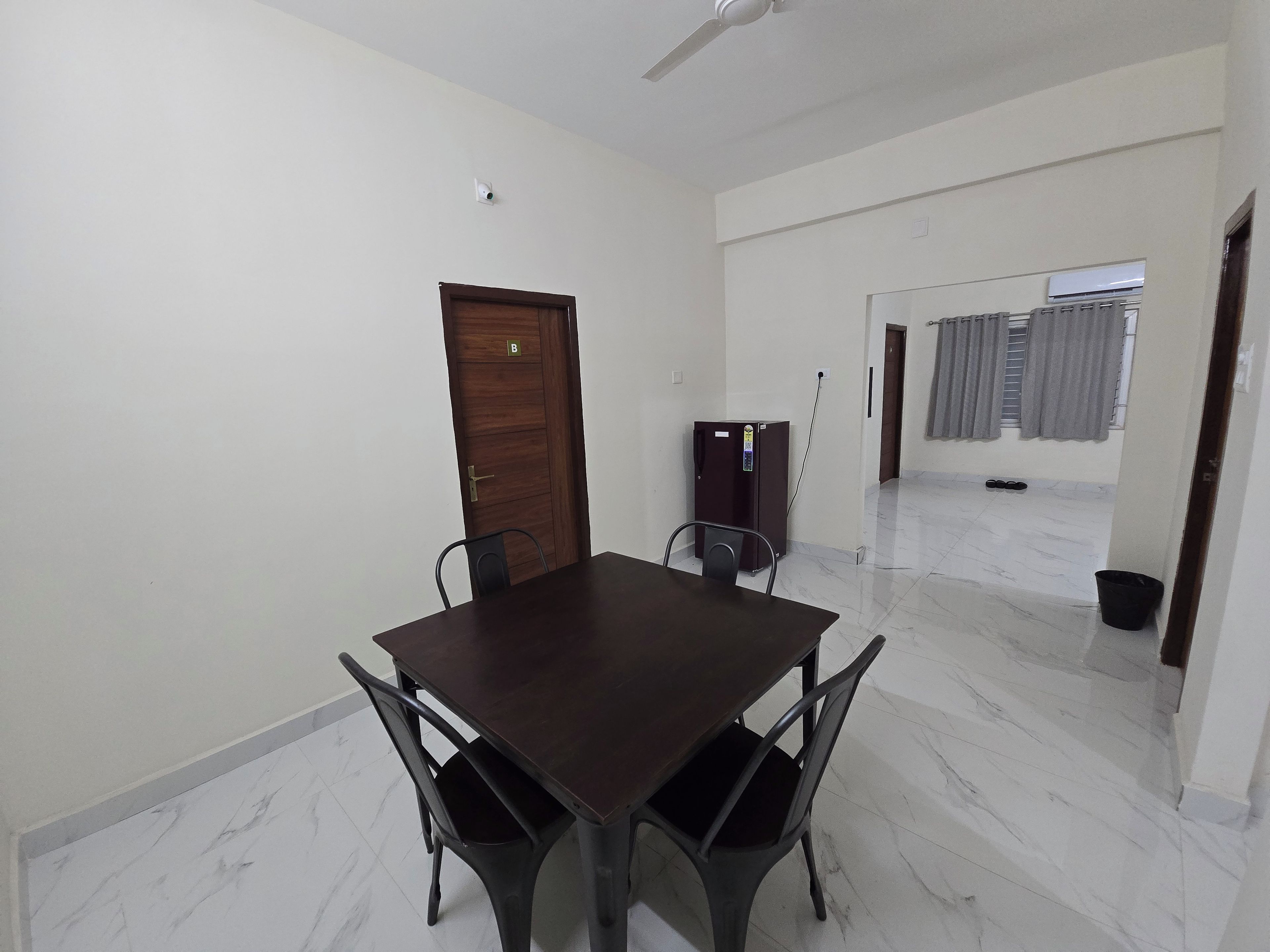 Una habitación con puerta de madera, un refrigerador, una mesa de comedor con sillas y un pasillo con piso de baldosas.