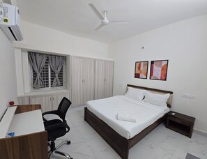 A hotel room with a bed, a desk, a chair, a ceiling fan, a window with curtains, and two framed pictures on the wall. - Manzl Collective Peepal  (Tada)
