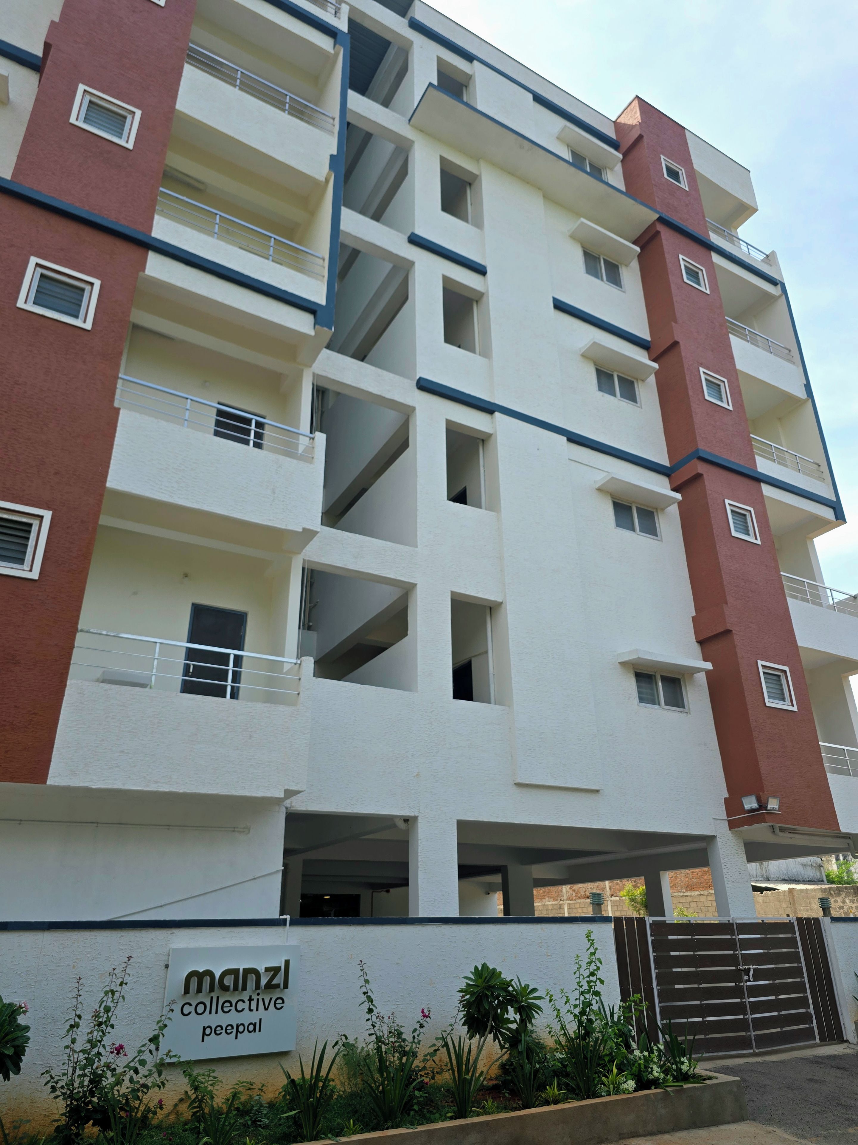A multi-story residential building with balconies, a gated entrance, and a sign indicating 'manzil collective peepal.'