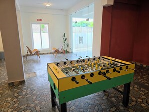A room with a foosball table, a fire exit sign, and a window with a view of greenery. - Manzl Collective Peepal  (Tada)
