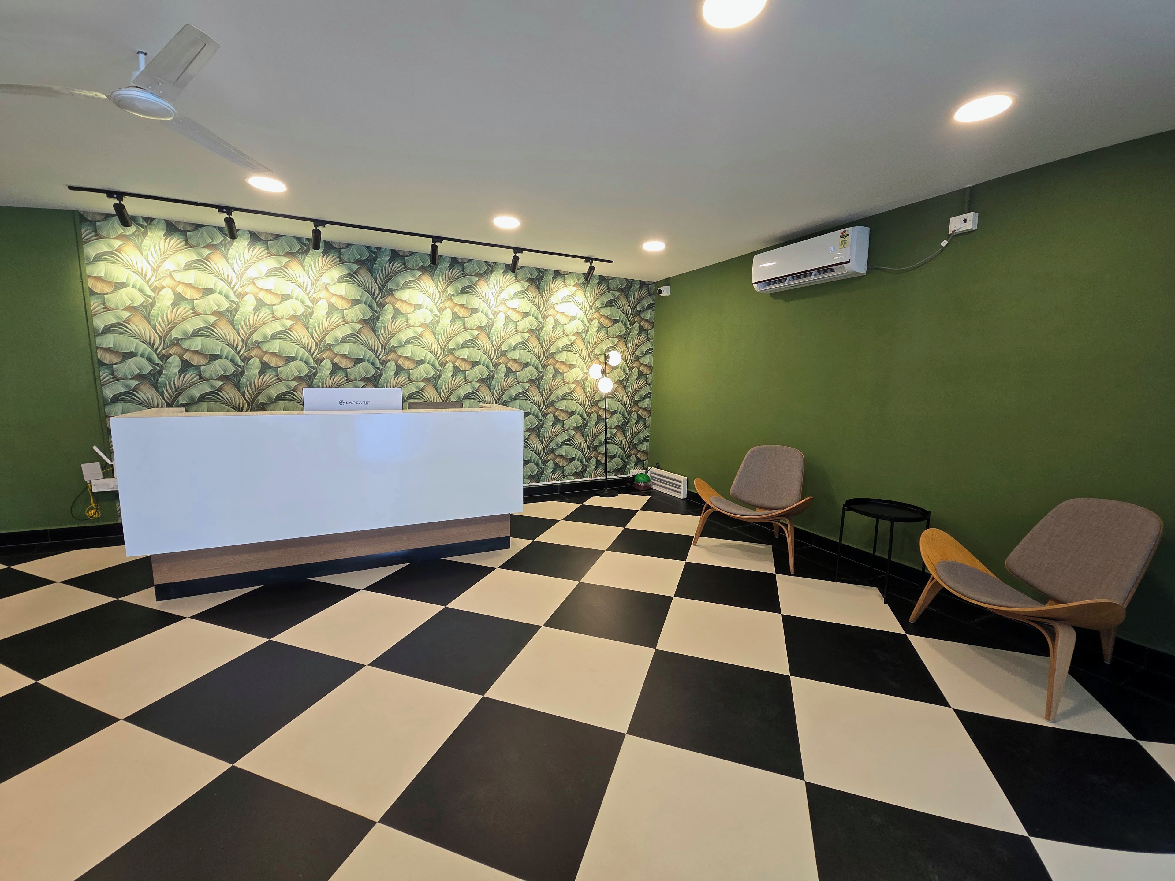 Un espace d’accueil avec un sol à carreaux, un comptoir de réception, deux chaises, un ventilateur de plafond, un éclairage encastré et un papier peint à motifs.