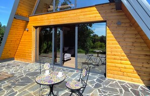 Eine Holzhütte mit einer Terrasse, ausgestattet mit Tisch und Stühlen, und einer Glasgleittür, die zu einem Sitzbereich im Freien führt.