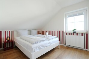 A bedroom with a white bed, striped wallpaper, a radiator, and a window with a plant. - Niendorf Residential Park, Apartment 7 - Niendorf Residential Park 13 (Timmendorfer Strand)