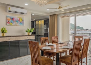A dining area with a table set for six, surrounded by wooden chairs, a ceiling fan, and a view of the sea through large windows. - ELIVAAS Signature Aura Sky 3 BHK Apt (Jaipur)