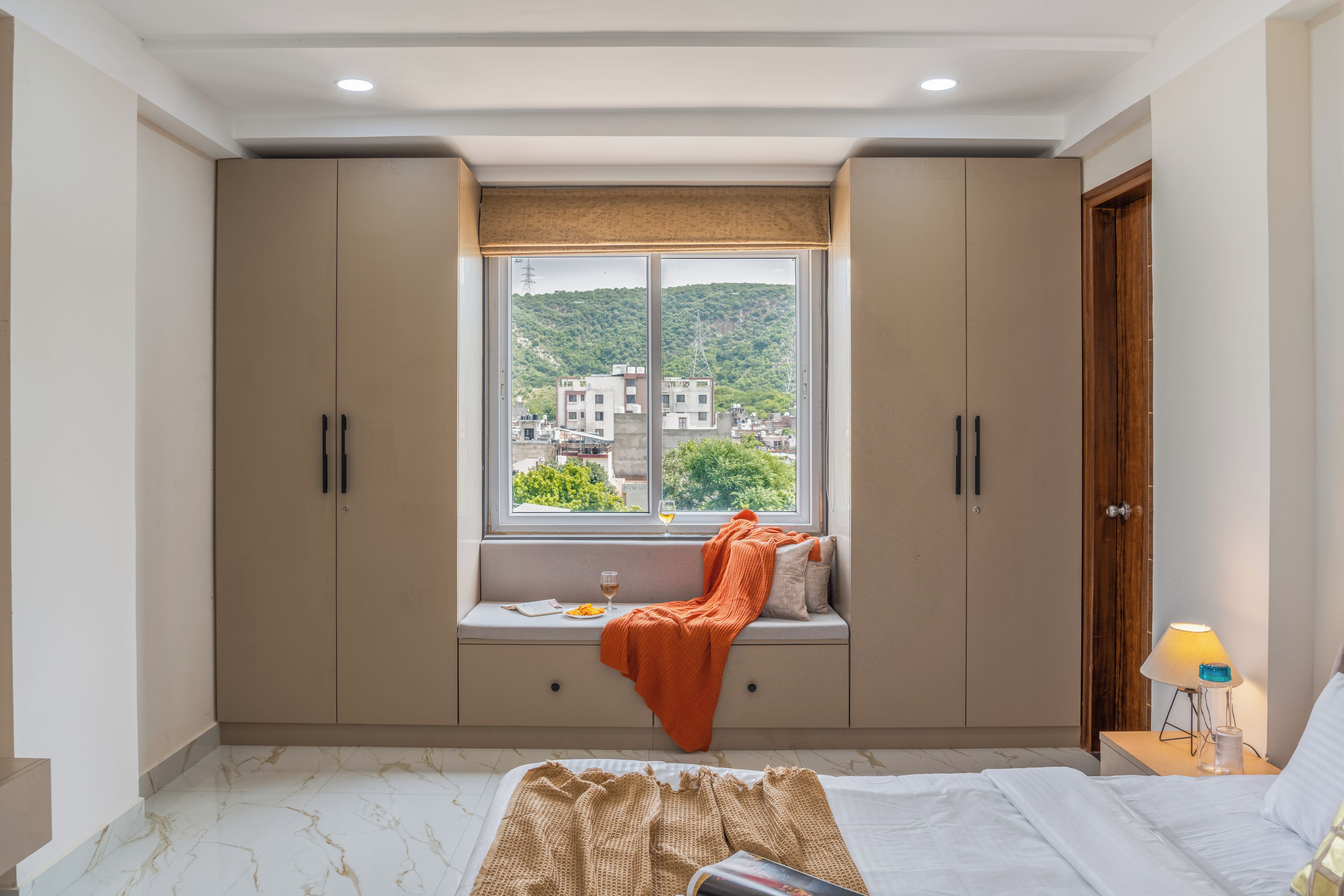 A bedroom with a large window, a bed, a bedside table with a lamp, and a wardrobe.