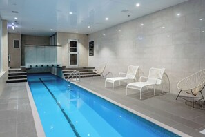Un espace piscine intérieur moderne, avec une piscine à l’eau d’un bleu limpide, des chaises longues blanches et un mur carrelé.