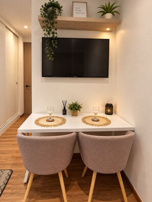 Ein Essbereich mit einem weißen Tisch, zwei Stühlen, einem an der Wand montierten Fernseher und einem Regal mit Pflanzen und einem gerahmten Zitat.