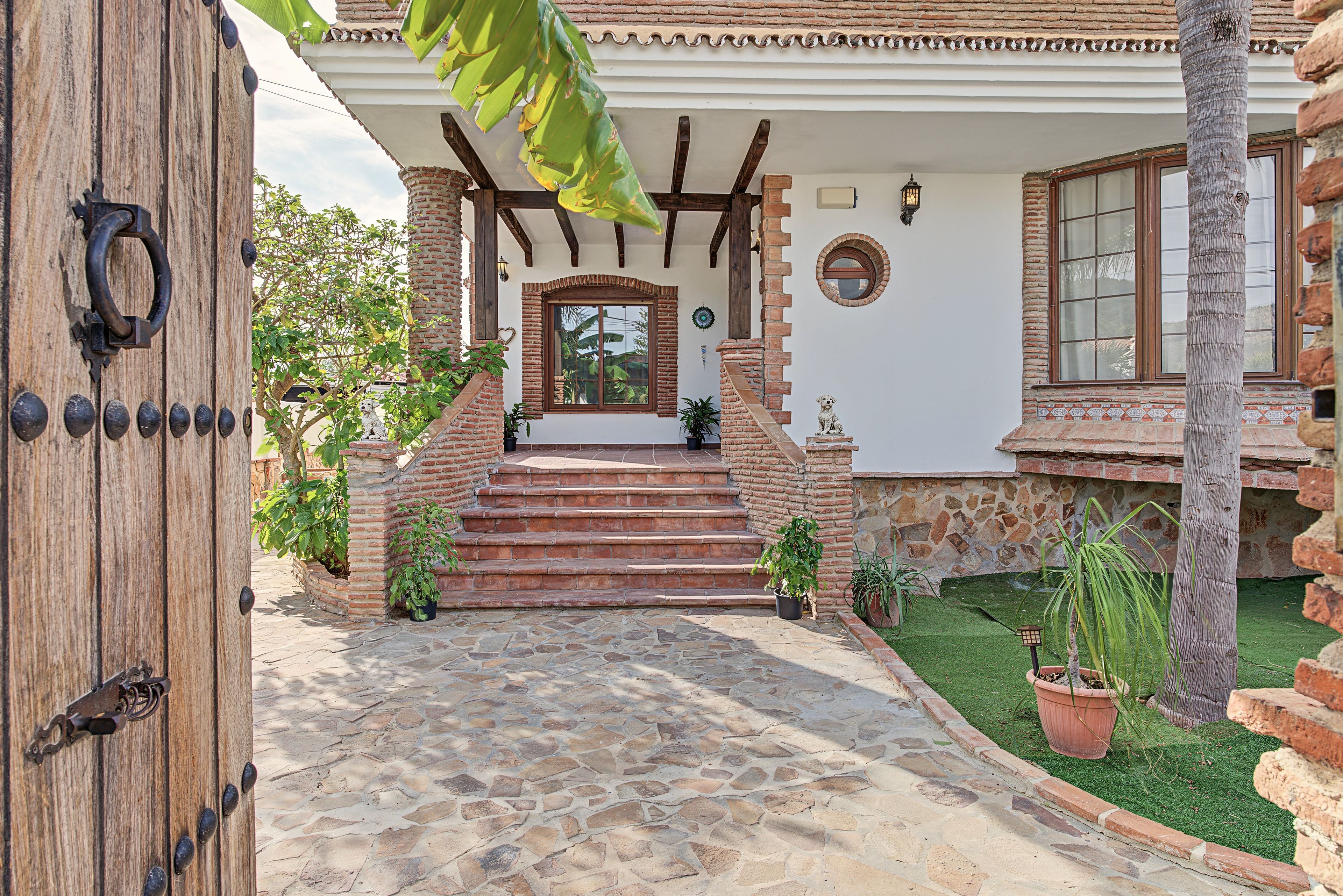 A house with a stone pathway leading to a wooden door, a brick staircase, and a small garden with potted plants.