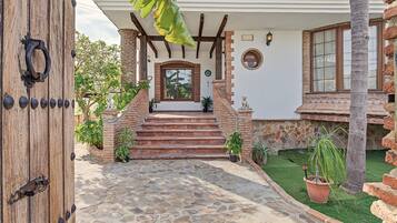 A house with a stone pathway leading to a wooden door, a brick staircase, and a small garden with potted plants.